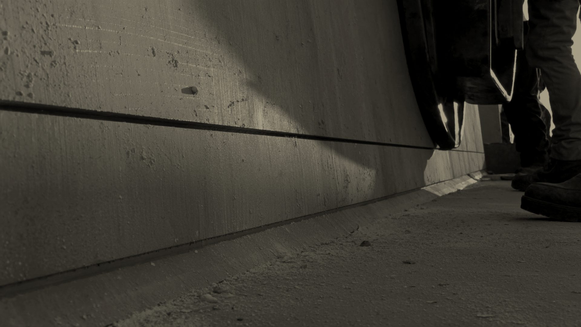 A straight control joint being cut across a fresh grey concrete slab with an early-entry saw, raking afternoon light showing the broom finish texture and fine concrete dust along the cut.