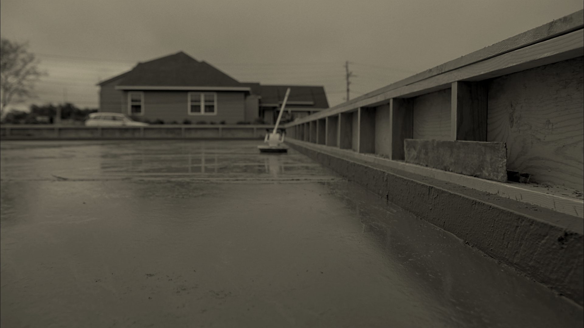 A freshly poured concrete driveway slab, still wet and grey, filling its timber forms end to end under an overcast sky, with a bull float lying across the far end